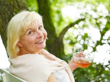 Eine ältere Dame sitzt auf einer Bank neben einem Baum und trinkt Tee.