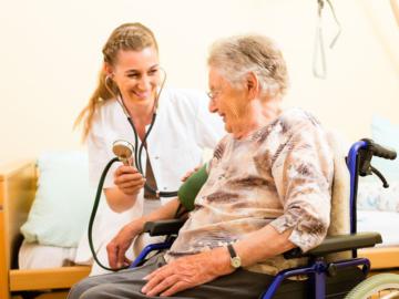 Eine Pflegerin misst einer älteren Frau im Rollstuhl den Blutdruck.
