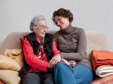 Ältere Frau mit Brille sitzt mit ihrer Betreuerin auf einer Couch - sie halten sich die Hände.