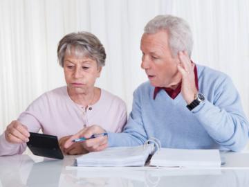 Ein älteres Paar sitzt vor einem Aktenordner mit einem Taschenrechner in der Hand.