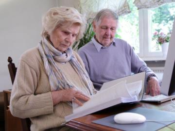 Ein älteres Paar sitzt vor einem Computer und blättert in einem Aktenordner.